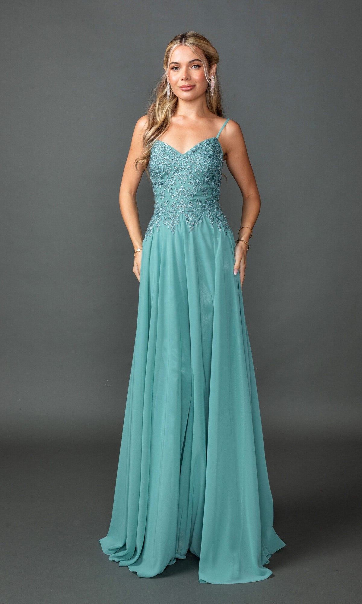 A woman stands against a gray background wearing the Dancing Queen 4542, a floor-length teal prom dress featuring spaghetti straps, a fitted lace embroidered bodice, and a flowing skirt.